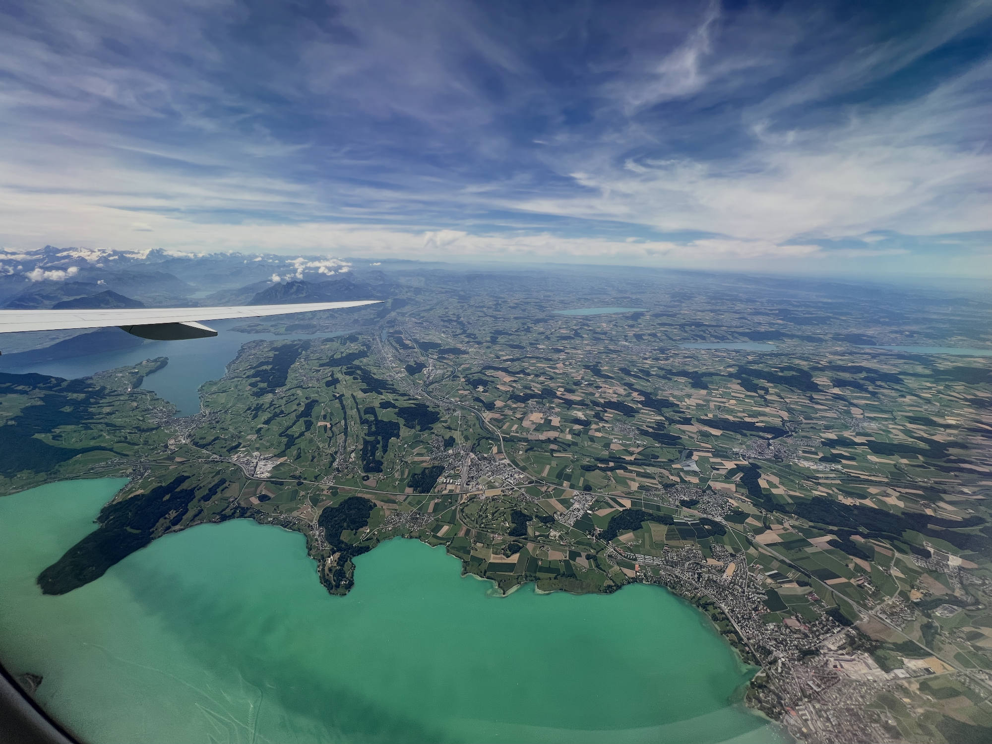 An aerial view taken from an airplane window shows a wingtip extending over a vast landscape of green fields, towns, and a bright turquoise lake. In the distance, snow-capped mountains rise beneath a blue sky filled with wispy white clouds.