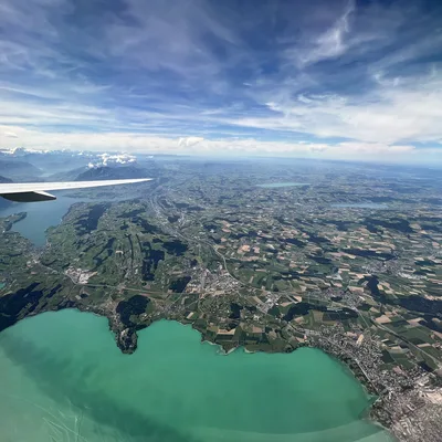 An aerial view taken from an airplane window shows a wingtip extending over a vast landscape of green fields, towns, and a bright turquoise lake. In the distance, snow-capped mountains rise beneath a blue sky filled with wispy white clouds.