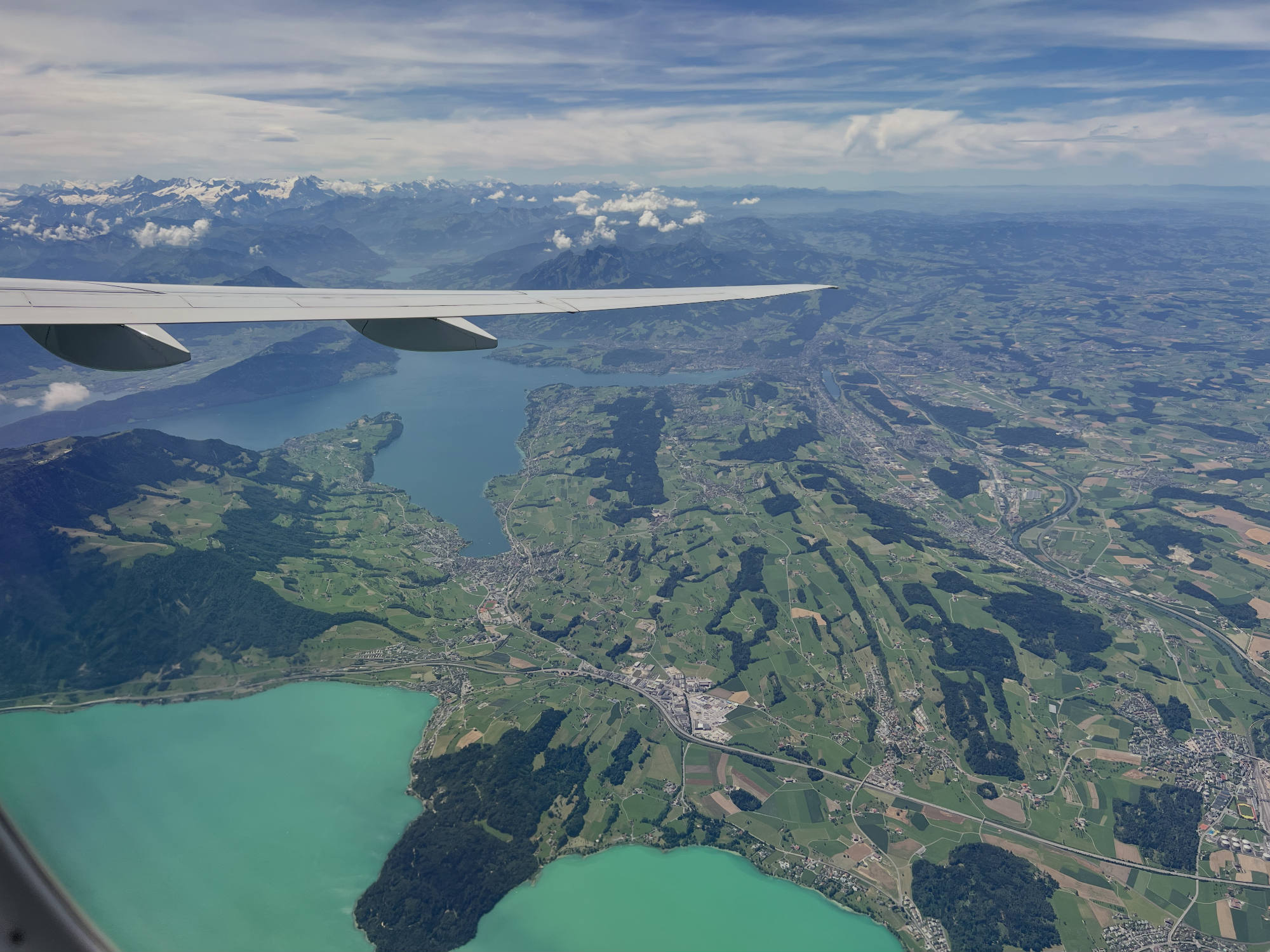 An aerial view from an airplane window shows the aircraft's wing extending over a landscape of turquoise lakes, green fields, and distant snow-capped mountains under a partly cloudy sky.