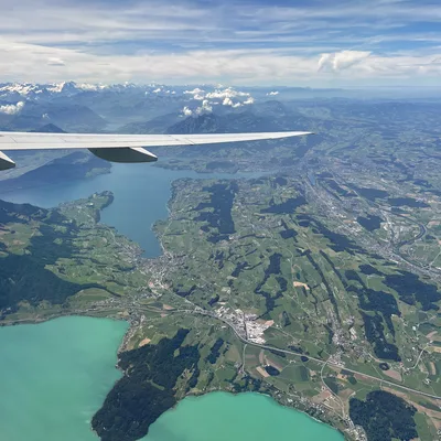An aerial view from an airplane window shows the aircraft's wing extending over a landscape of turquoise lakes, green fields, and distant snow-capped mountains under a partly cloudy sky.