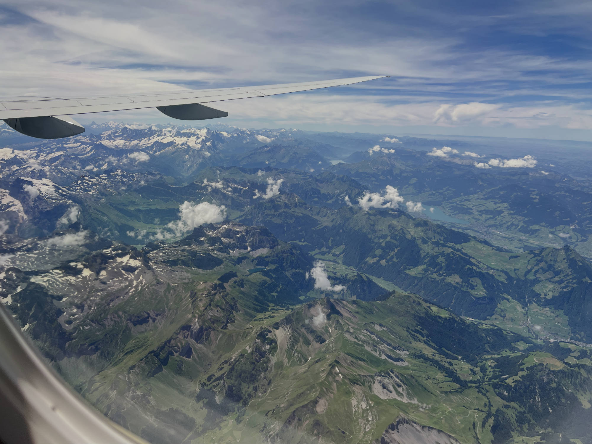 An aerial view taken from an airplane window shows the aircraft's wing extending over a vast mountain range with green valleys, snow-capped peaks, and scattered clouds. A blue sky with wispy clouds stretches across the horizon above the mountainous landscape.
