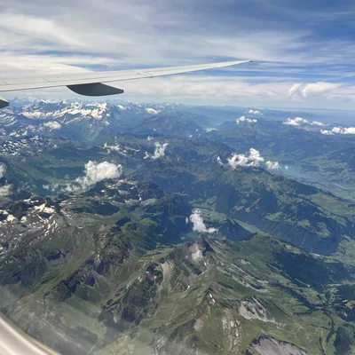 An aerial view taken from an airplane window shows the aircraft's wing extending over a vast mountain range with green valleys, snow-capped peaks, and scattered clouds. A blue sky with wispy clouds stretches across the horizon above the mountainous landscape.