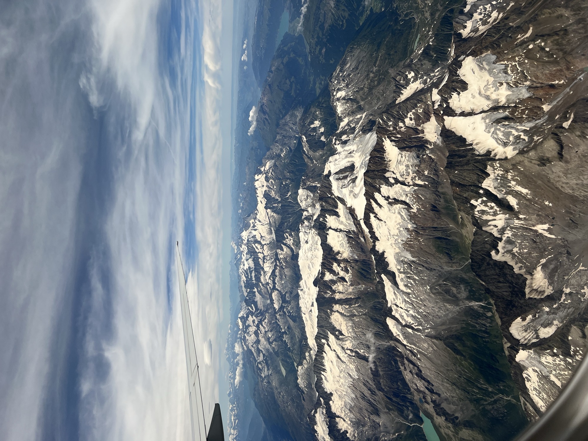 An aerial view from an airplane window captures a vast, snow-capped mountain range under a partly cloudy blue sky, with the tip of the plane's wing visible in the upper left. The rugged peaks and valleys are covered in patches of snow and ice, with a small turquoise lake visible in the lower left.