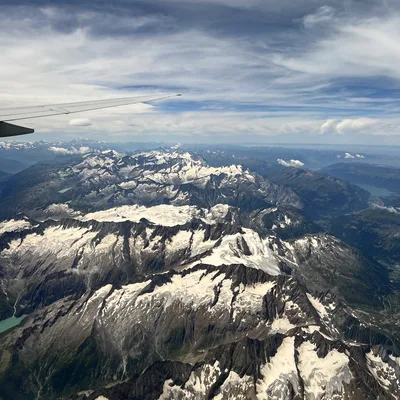 An aerial view from an airplane window captures a vast, snow-capped mountain range under a partly cloudy blue sky, with the tip of the plane's wing visible in the upper left. The rugged peaks and valleys are covered in patches of snow and ice, with a small turquoise lake visible in the lower left.