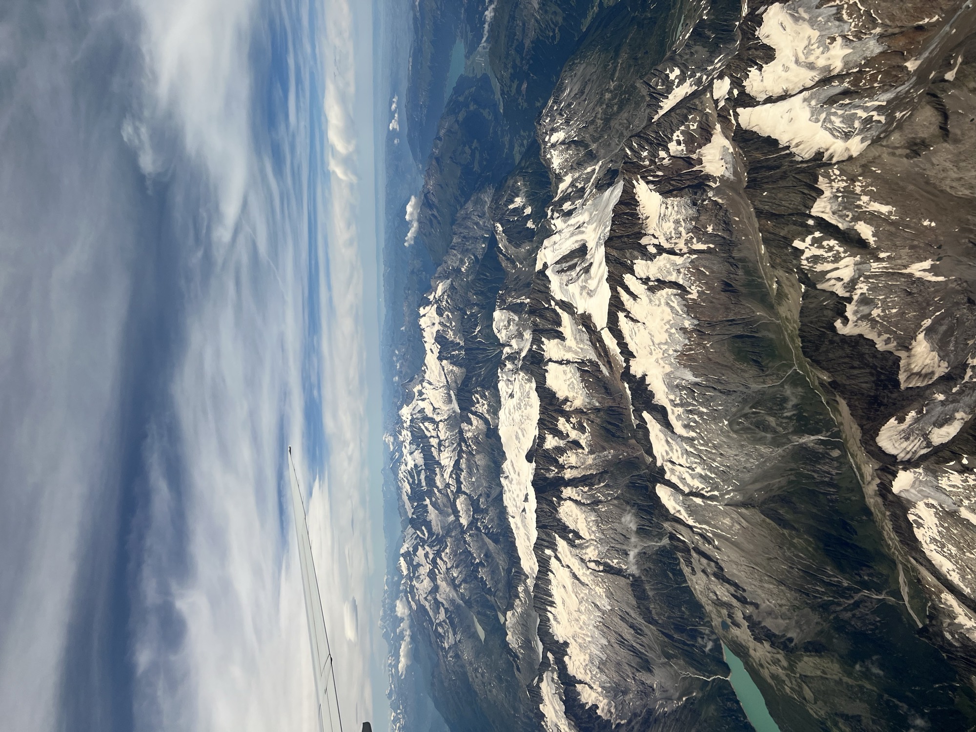 An aerial view taken from an airplane window shows snow-capped mountain peaks under a cloudy blue sky. The tip of the airplane's wing is visible in the upper left corner of the frame.