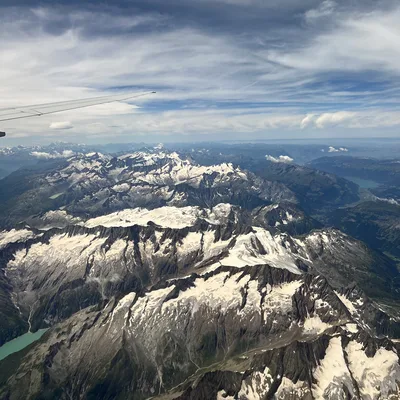 An aerial view taken from an airplane window shows snow-capped mountain peaks under a cloudy blue sky. The tip of the airplane's wing is visible in the upper left corner of the frame.