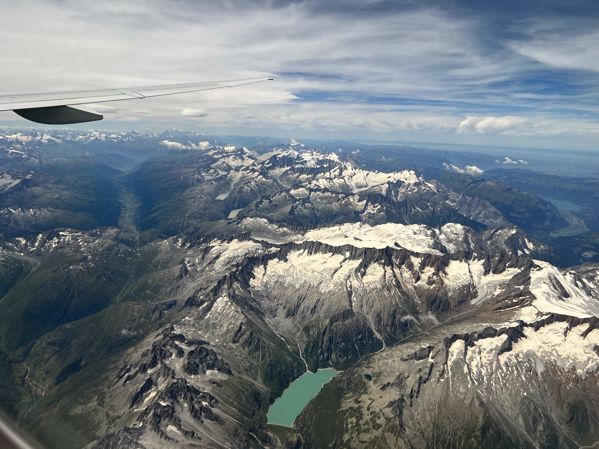 An aerial view taken from an airplane window shows a vast, snow-capped mountain range with a turquoise alpine lake nestled in a valley below. The wing of the airplane is visible in the upper left corner against a partly cloudy blue sky.