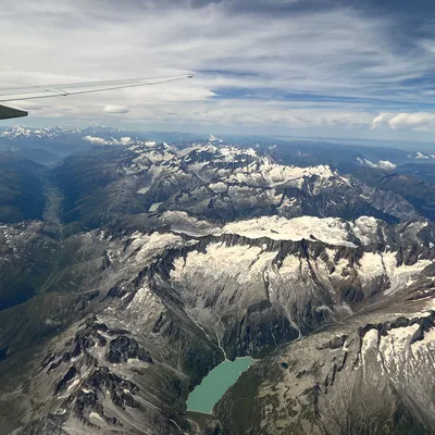 An aerial view taken from an airplane window shows a vast, snow-capped mountain range with a turquoise alpine lake nestled in a valley below. The wing of the airplane is visible in the upper left corner against a partly cloudy blue sky.
