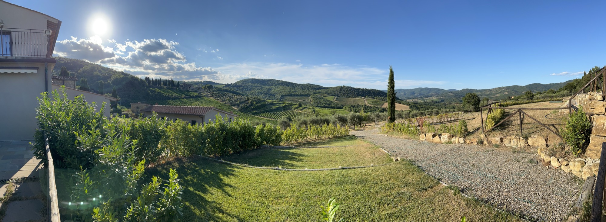 This panoramic photograph captures a sunny countryside landscape featuring rolling green hills, vineyards, and a gravel path leading through a grassy yard. A building with a tiled roof sits on the left, set against a bright blue sky with scattered clouds and a shining sun.