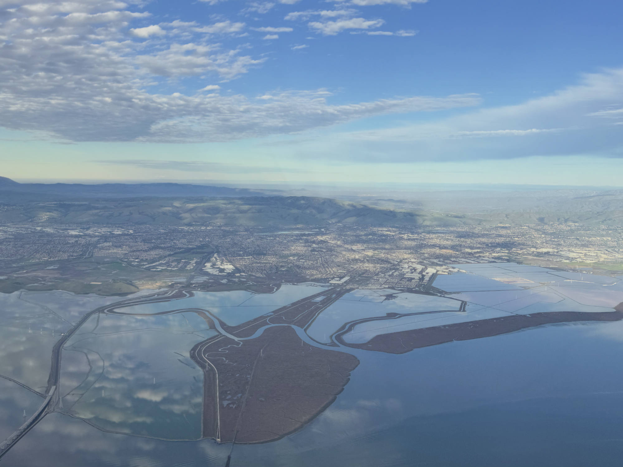 An aerial view captures a coastal landscape featuring salt ponds, wetlands, and a city sprawling toward distant hills under a partly cloudy blue sky. The sky and clouds are reflected in the calm, flat waters of the bay and ponds below.