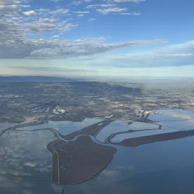 An aerial view captures a coastal landscape featuring salt ponds, wetlands, and a city sprawling toward distant hills under a partly cloudy blue sky. The sky and clouds are reflected in the calm, flat waters of the bay and ponds below.