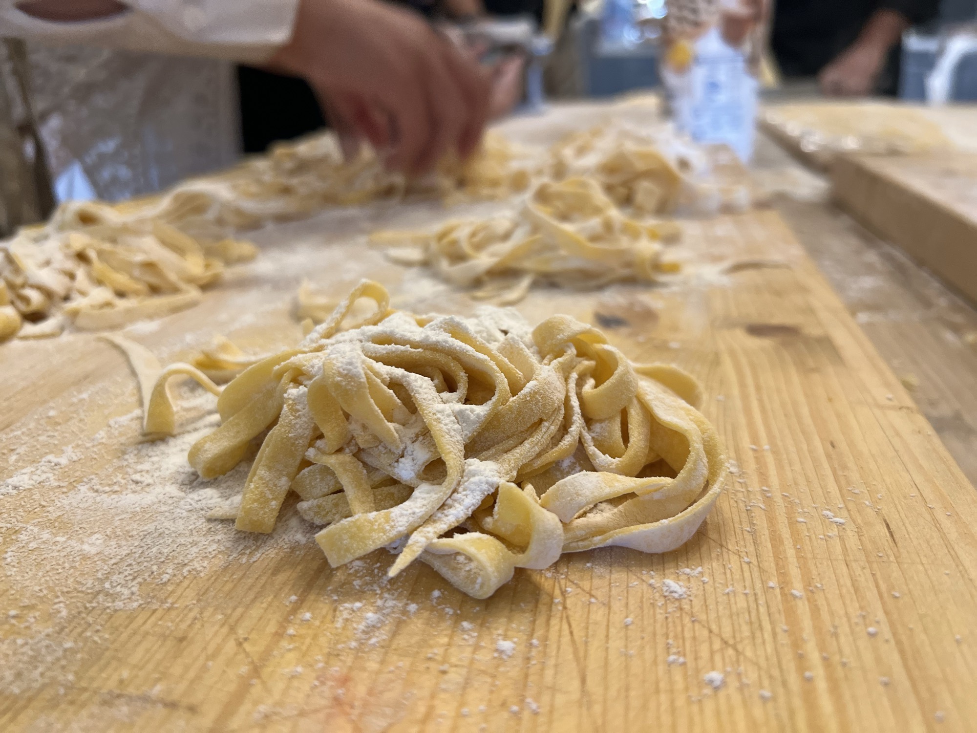 A close-up shot shows freshly made, flour-dusted pasta noodles resting on a wooden work surface. In the blurred background, people are actively preparing more pasta in what appears to be a kitchen or cooking class setting.