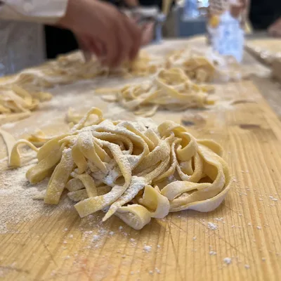 A close-up shot shows freshly made, flour-dusted pasta noodles resting on a wooden work surface. In the blurred background, people are actively preparing more pasta in what appears to be a kitchen or cooking class setting.