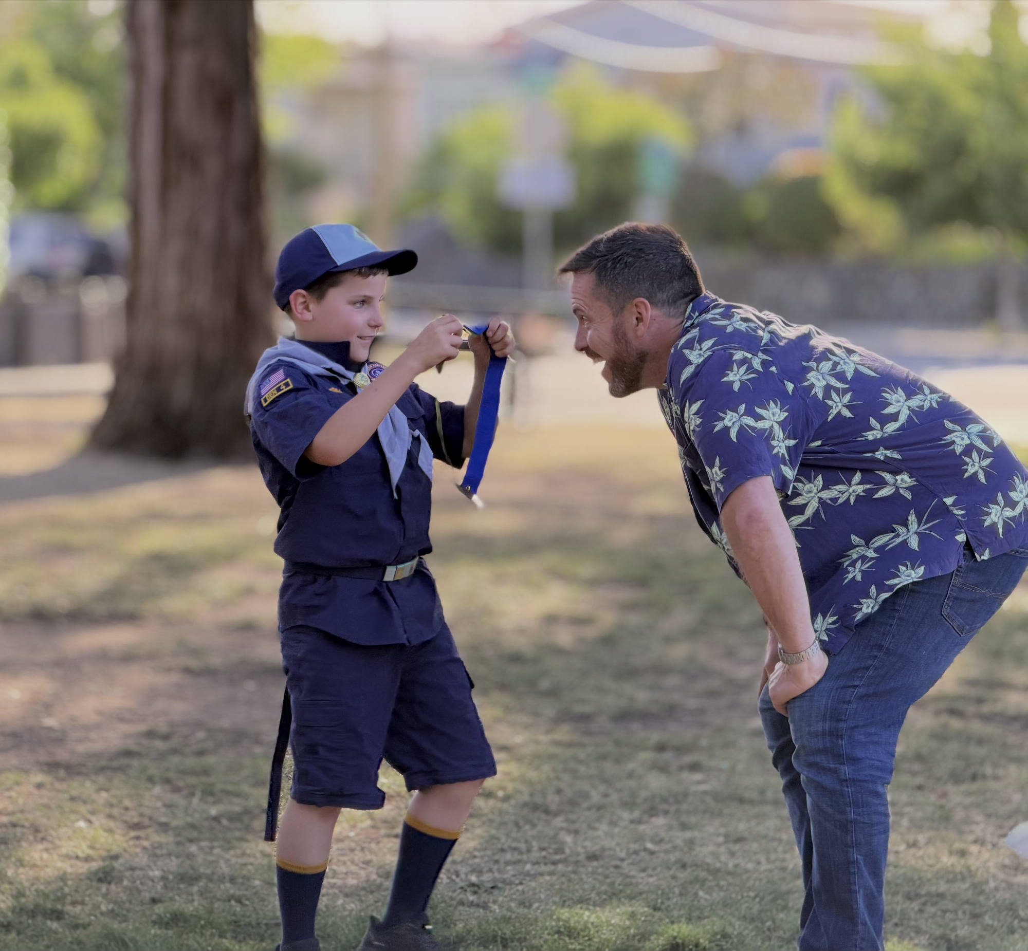 In an outdoor park setting, a young boy in a Cub Scout uniform proudly shows a blue ribbon and medal to a smiling man leaning in to look.