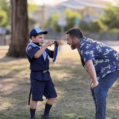 In an outdoor park setting, a young boy in a Cub Scout uniform proudly shows a blue ribbon and medal to a smiling man leaning in to look.