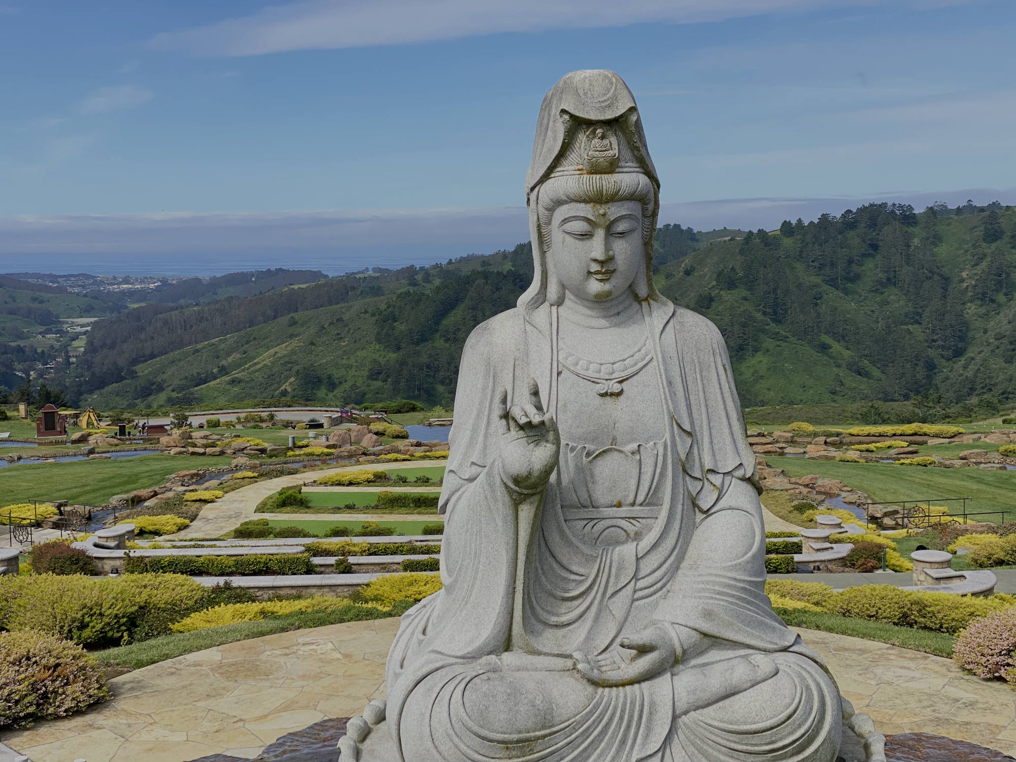 A large stone statue of Guanyin sits in a meditative pose on a paved overlook. In the background, a landscaped garden with yellow flowers and green lawns slopes down toward forested hills under a blue sky.