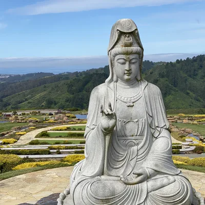 A large stone statue of Guanyin sits in a meditative pose on a paved overlook. In the background, a landscaped garden with yellow flowers and green lawns slopes down toward forested hills under a blue sky.