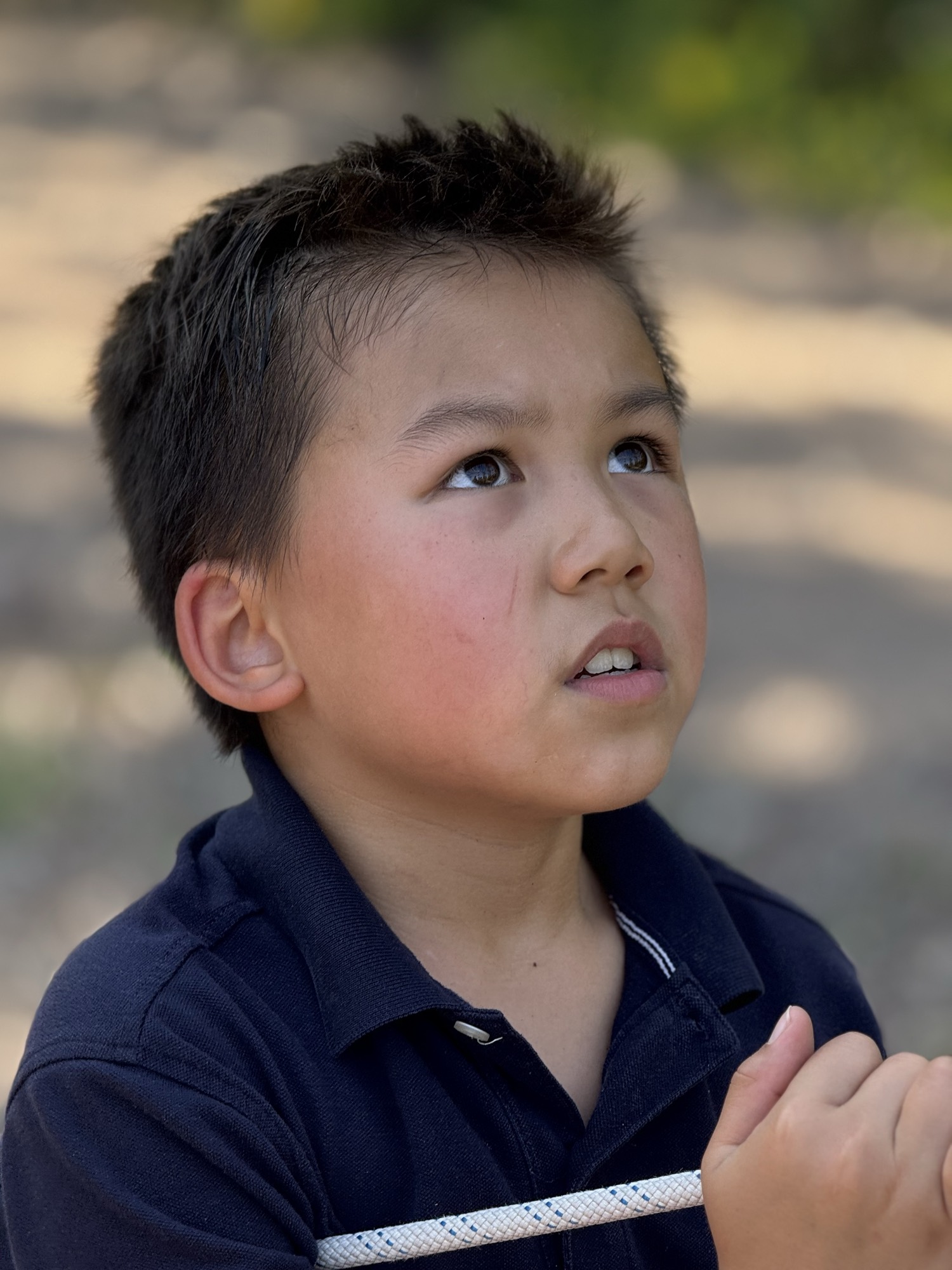 A close-up portrait shows a young boy in a dark polo shirt looking upward with a focused expression while holding a white rope. The background is a softly blurred outdoor setting with natural light.