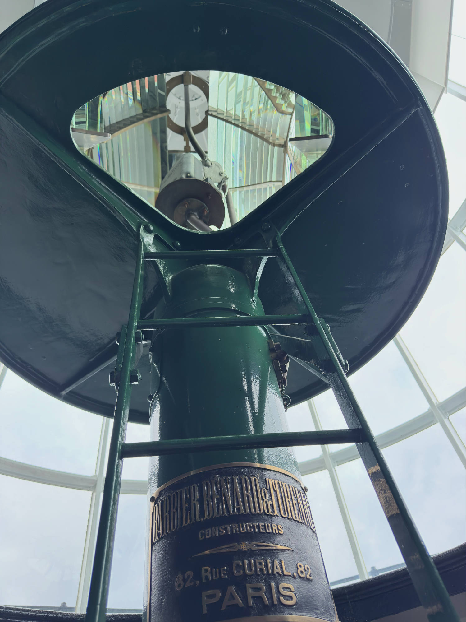 This low-angle shot captures the green metal support structure and ladder of a historic lighthouse lens, featuring a manufacturer's plaque that reads "Barbier, Benard & Turenne, Constructeurs, 82, Rue Curial, 82, Paris." The mechanism is housed indoors behind large glass windows, with the Fresnel lens visible through an opening at the top.