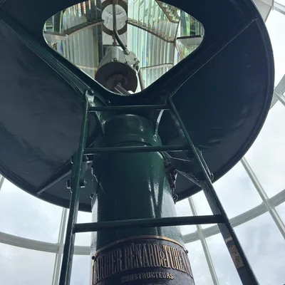 This low-angle shot captures the green metal support structure and ladder of a historic lighthouse lens, featuring a manufacturer's plaque that reads "Barbier, Benard & Turenne, Constructeurs, 82, Rue Curial, 82, Paris." The mechanism is housed indoors behind large glass windows, with the Fresnel lens visible through an opening at the top.