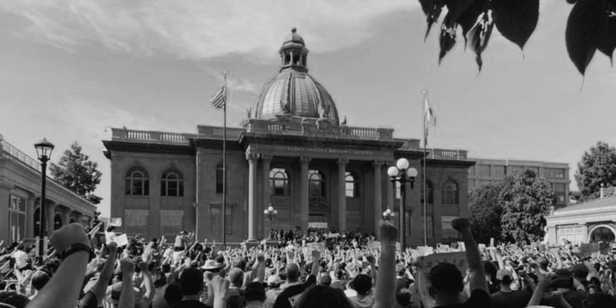 A large crowd of people is gathered outside a domed government building, raising their fists in protest. This black-and-white photograph captures the demonstration from behind the crowd, looking toward the building's entrance.