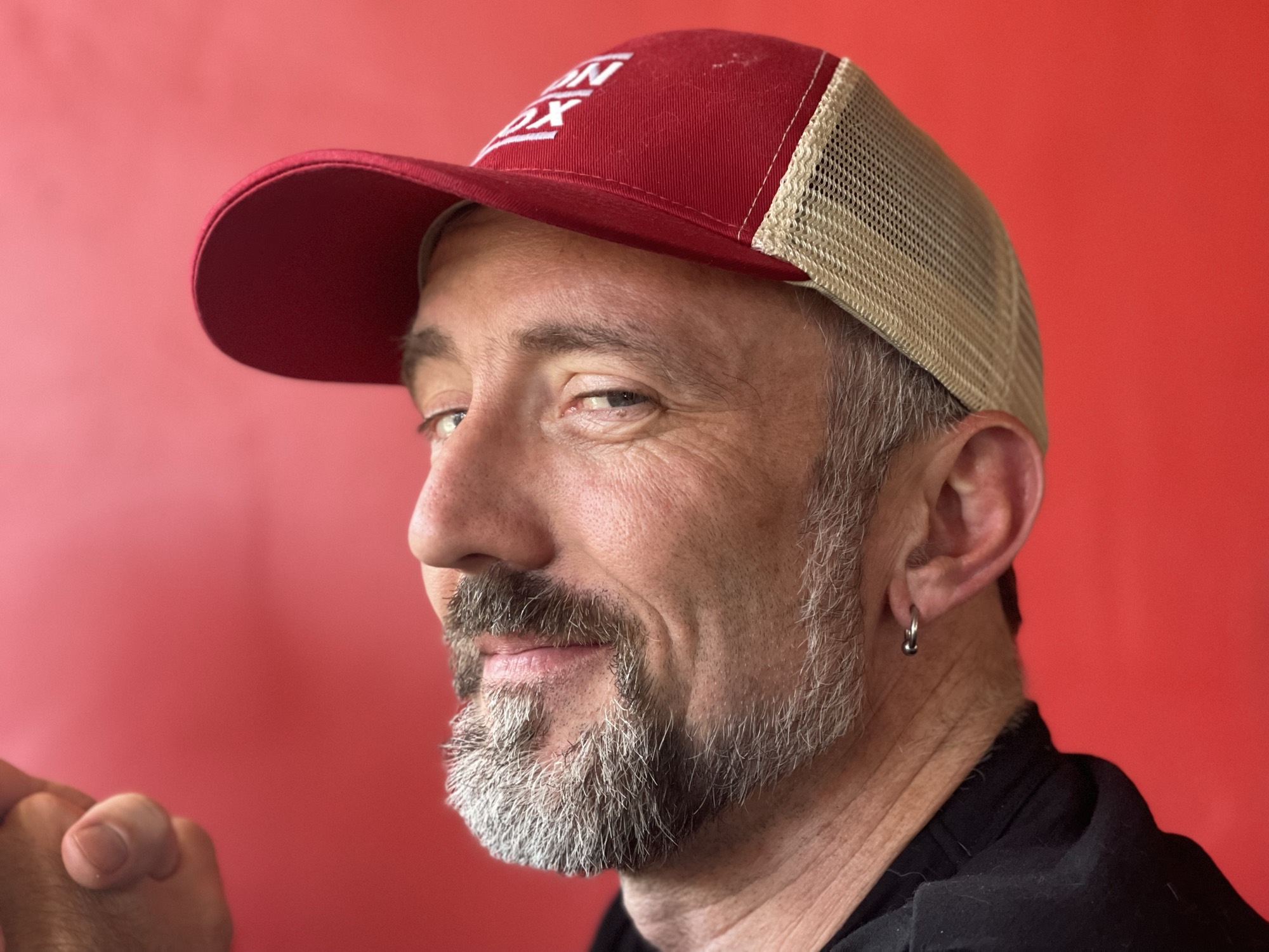 A close-up portrait of a smiling man with a grey-flecked beard, wearing a red and beige trucker hat and a small hoop earring. He is looking toward the camera in profile against a solid red background.