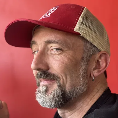 A close-up portrait of a smiling man with a grey-flecked beard, wearing a red and beige trucker hat and a small hoop earring. He is looking toward the camera in profile against a solid red background.