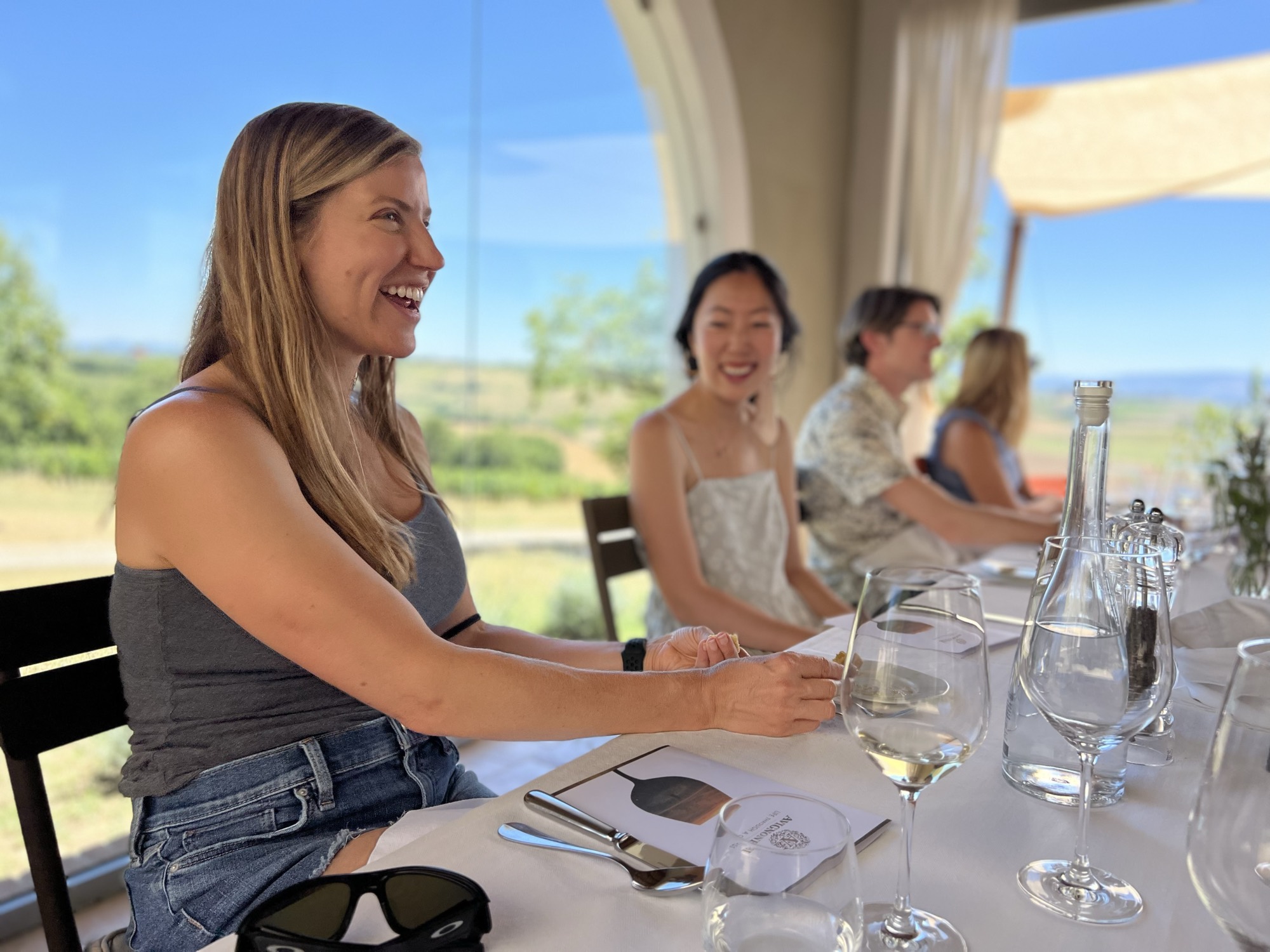 A group of people are seated at a long, white-clothed table for an outdoor dining or wine tasting experience under a covered patio. The foreground features a smiling woman in a grey tank top and denim shorts, with wine glasses, water bottles, and menus arranged on the table overlooking a scenic, sunny landscape.