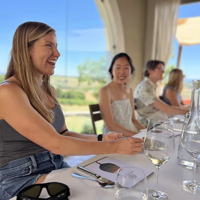 A group of people are seated at a long, white-clothed table for an outdoor dining or wine tasting experience under a covered patio. The foreground features a smiling woman in a grey tank top and denim shorts, with wine glasses, water bottles, and menus arranged on the table overlooking a scenic, sunny landscape.