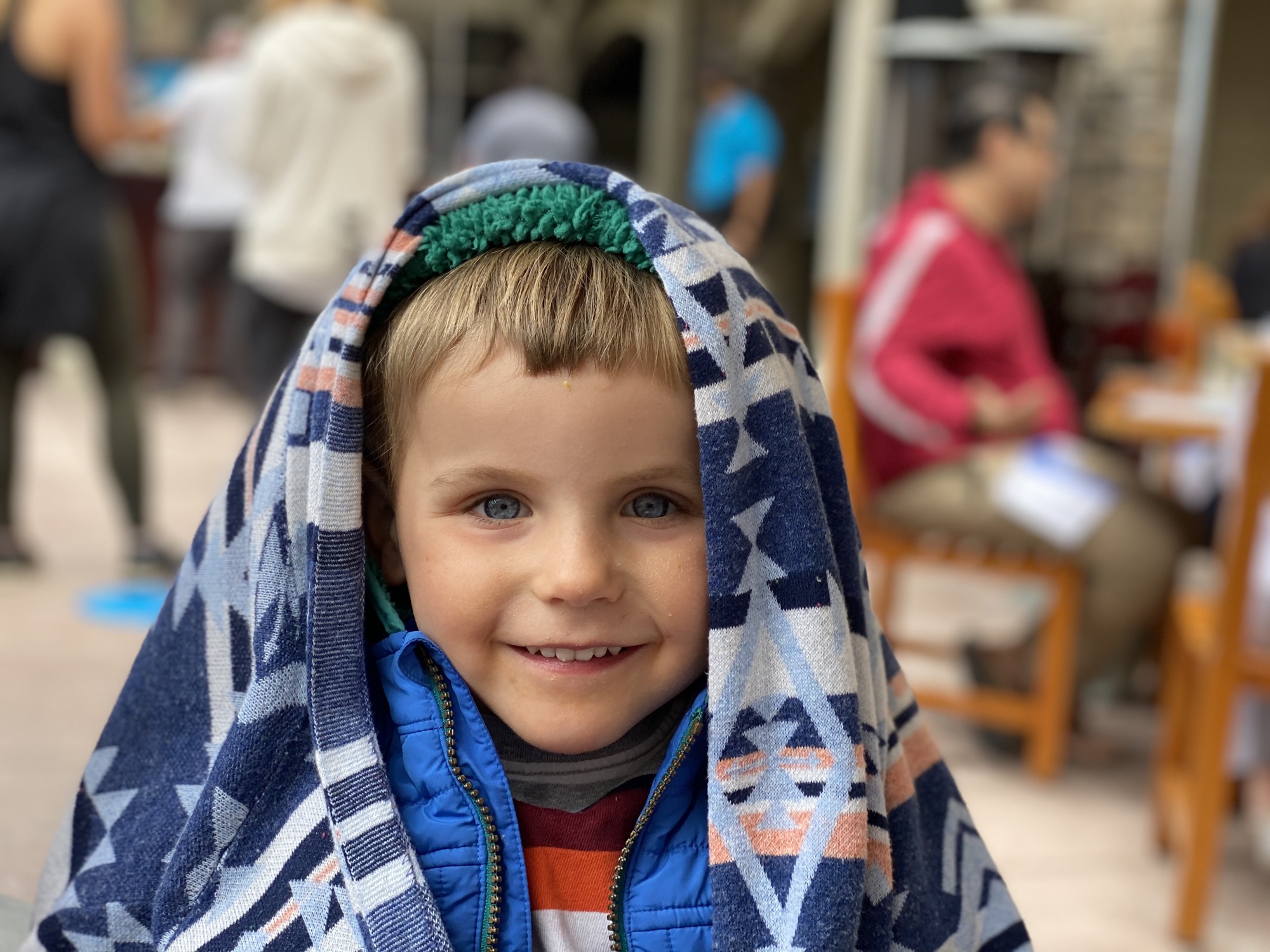 A young child with blue eyes smiles at the camera while wearing a blue jacket and a patterned blue and white blanket draped over their head. The child is outdoors in a bustling, blurred public area with other people and outdoor seating in the background.