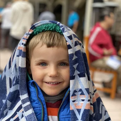 A young child with blue eyes smiles at the camera while wearing a blue jacket and a patterned blue and white blanket draped over their head. The child is outdoors in a bustling, blurred public area with other people and outdoor seating in the background.