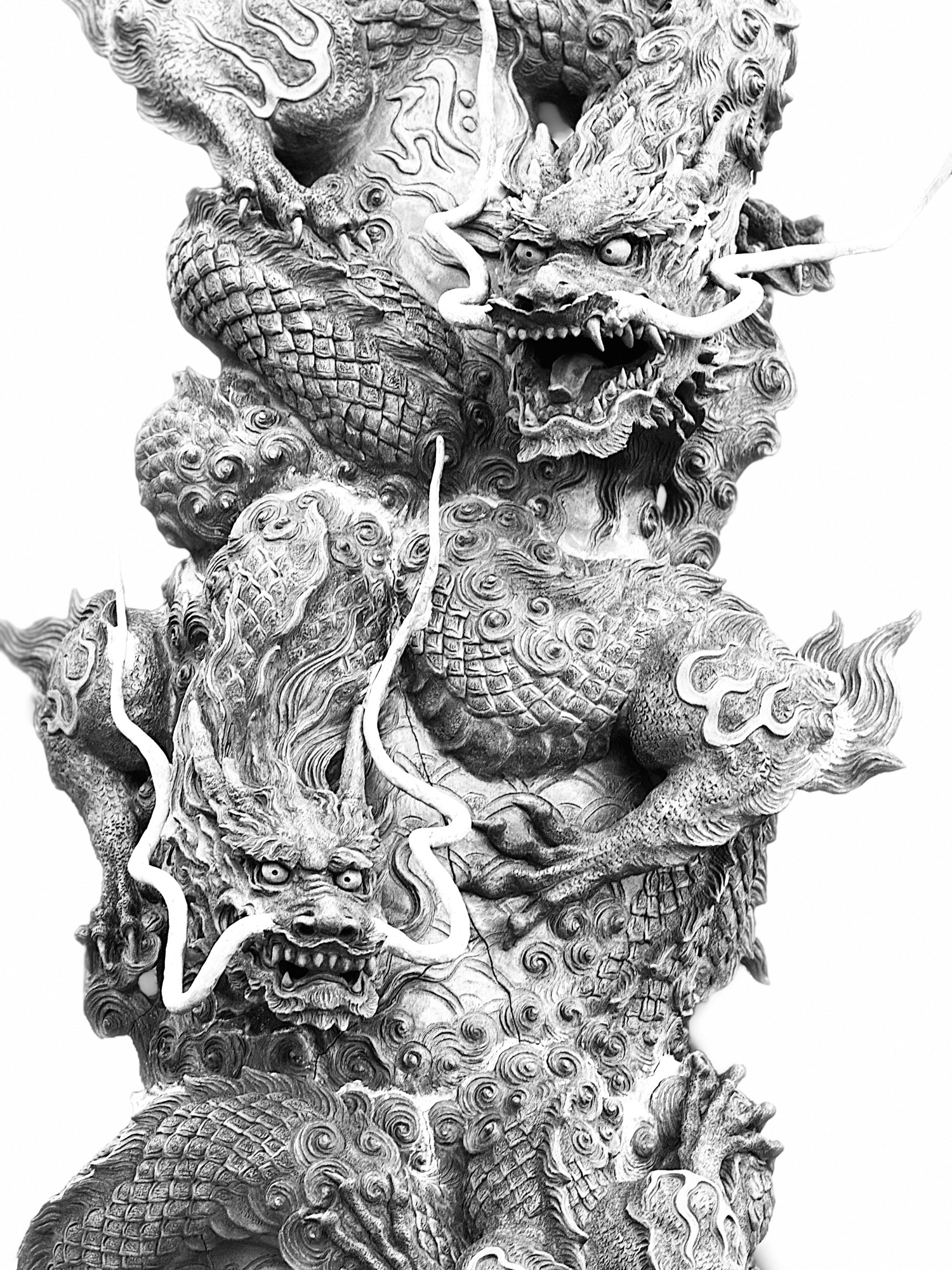 This black and white close-up photograph features a highly detailed, ornate stone pillar carved with two Chinese dragons. The sculpture is set against a plain white background, highlighting the intricate textures of the scales, whiskers, and swirling clouds.