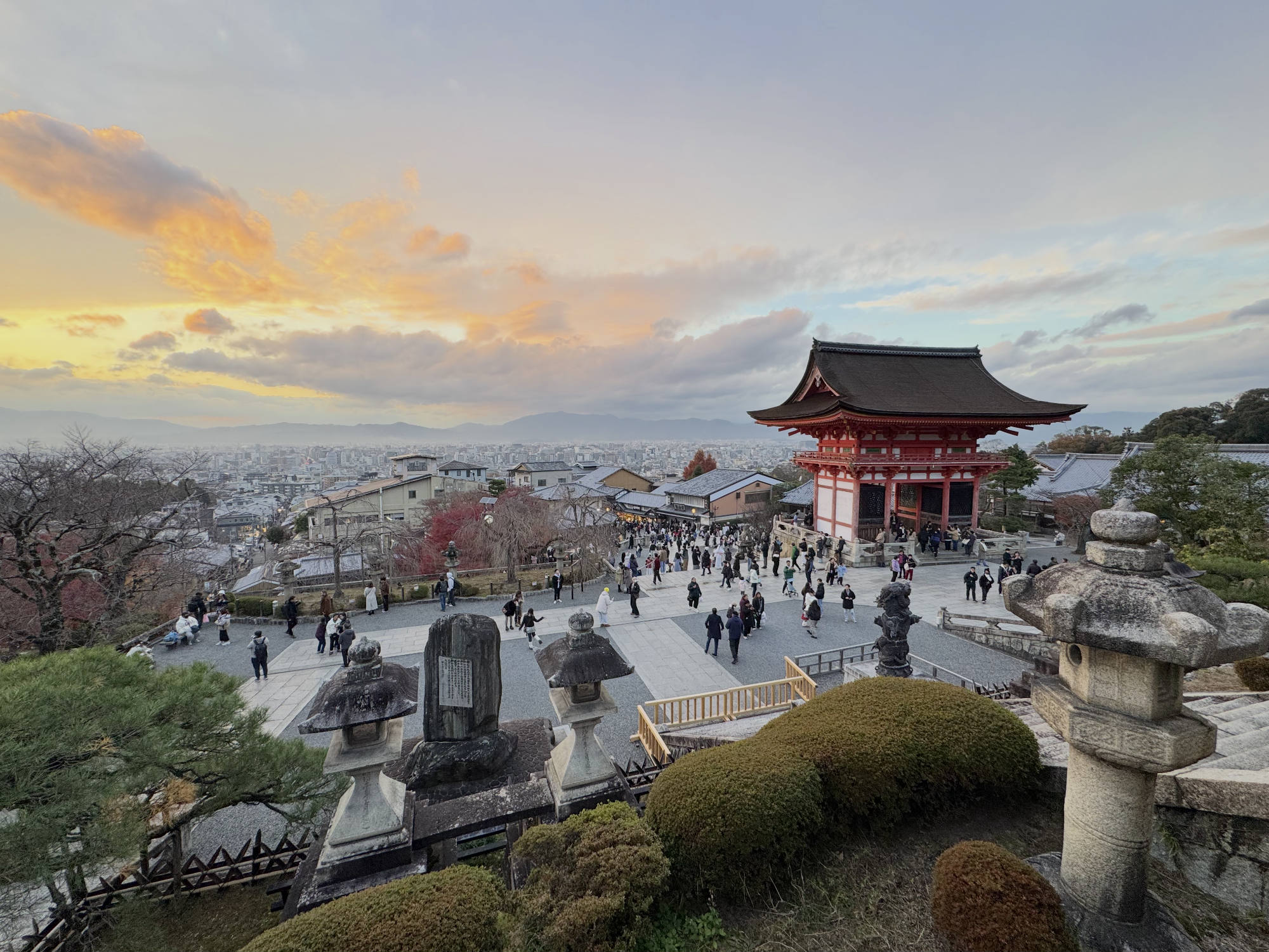 A high-angle shot captures numerous visitors walking around the grounds of a traditional Japanese temple complex, featuring a prominent red pagoda-style gate, overlooking a city in the distance. The scene is set against a dramatic sunset sky with orange-tinged clouds, framed by stone lanterns and manicured shrubbery in the foreground.