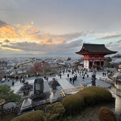 A high-angle shot captures numerous visitors walking around the grounds of a traditional Japanese temple complex, featuring a prominent red pagoda-style gate, overlooking a city in the distance. The scene is set against a dramatic sunset sky with orange-tinged clouds, framed by stone lanterns and manicured shrubbery in the foreground.