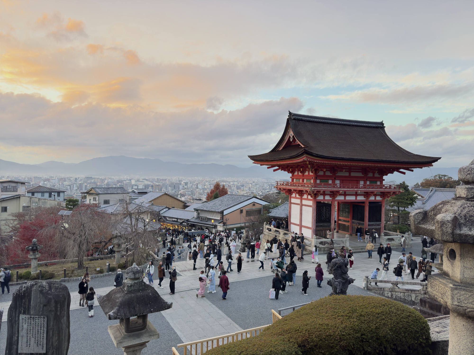 A large crowd of people walks around the stone courtyard of a traditional Japanese temple complex, which features a prominent red and white gatehouse. In the background, a sprawling city stretches toward distant mountains under a dramatic sunset sky.