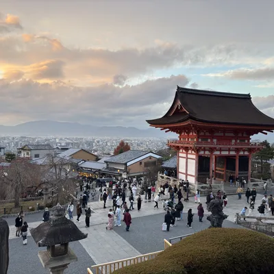 A large crowd of people walks around the stone courtyard of a traditional Japanese temple complex, which features a prominent red and white gatehouse. In the background, a sprawling city stretches toward distant mountains under a dramatic sunset sky.