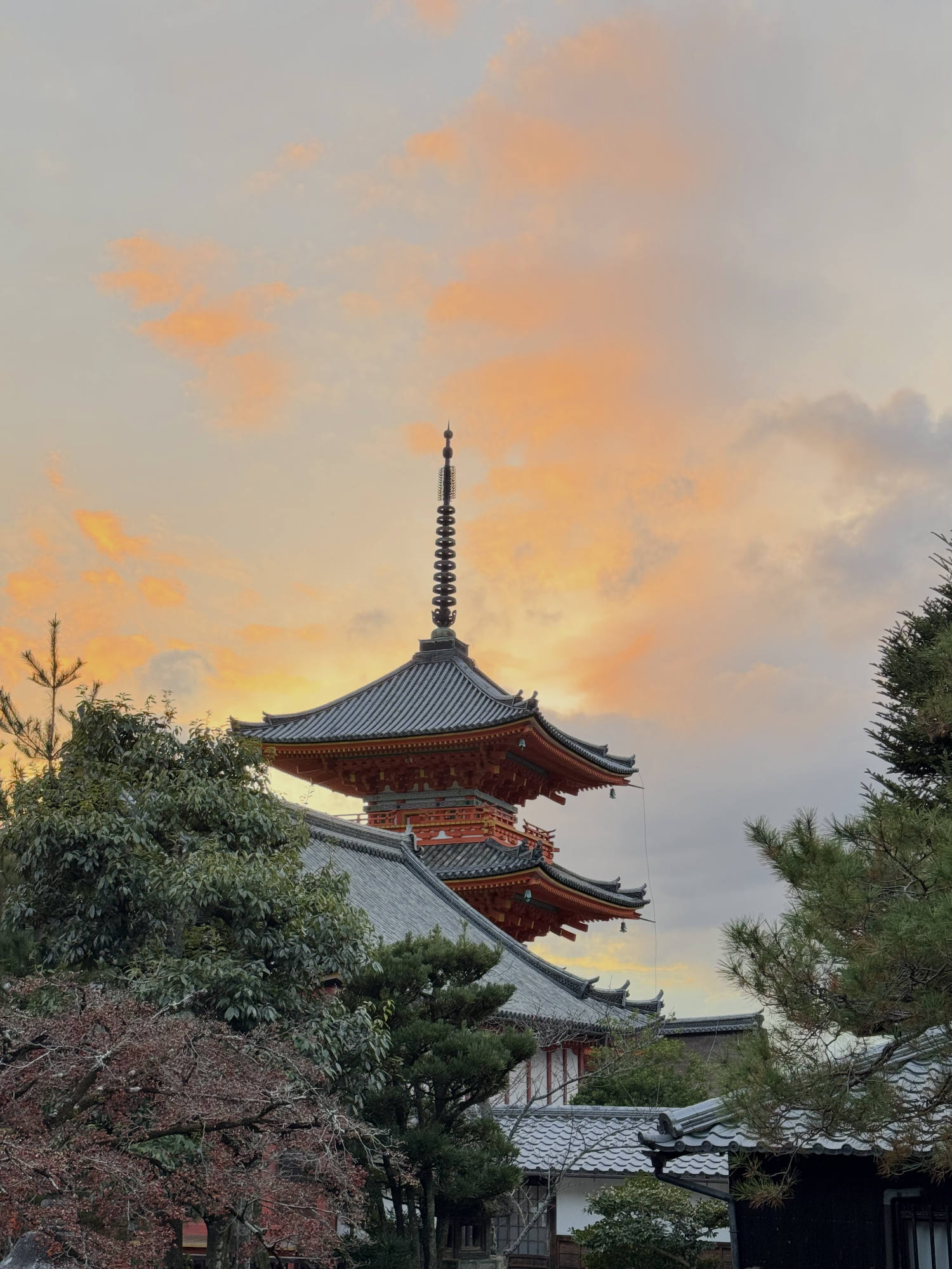 A multi-tiered Japanese pagoda with a red and black exterior rises above surrounding temple roofs and green trees. The scene is set against a dramatic sky filled with clouds illuminated by a golden sunset.