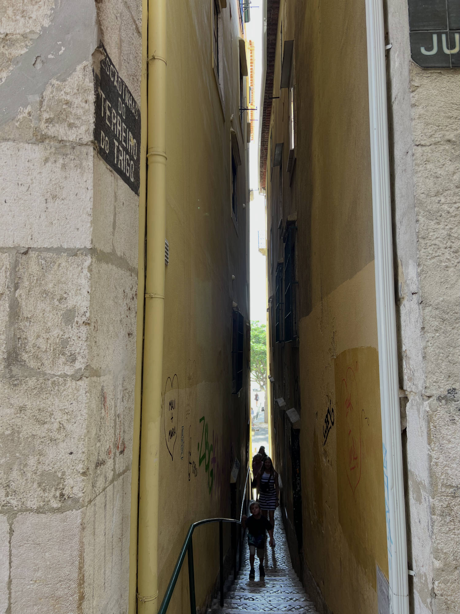 This eye-level shot captures a very narrow, cobblestone alleyway between two tall, yellow-painted buildings in Lisbon, Portugal, as indicated by the street sign reading "Escadinhas do Terreiro do Trigo." In the distance, a few pedestrians are walking up the stairs of the alleyway toward the camera.