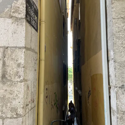 This eye-level shot captures a very narrow, cobblestone alleyway between two tall, yellow-painted buildings in Lisbon, Portugal, as indicated by the street sign reading "Escadinhas do Terreiro do Trigo." In the distance, a few pedestrians are walking up the stairs of the alleyway toward the camera.