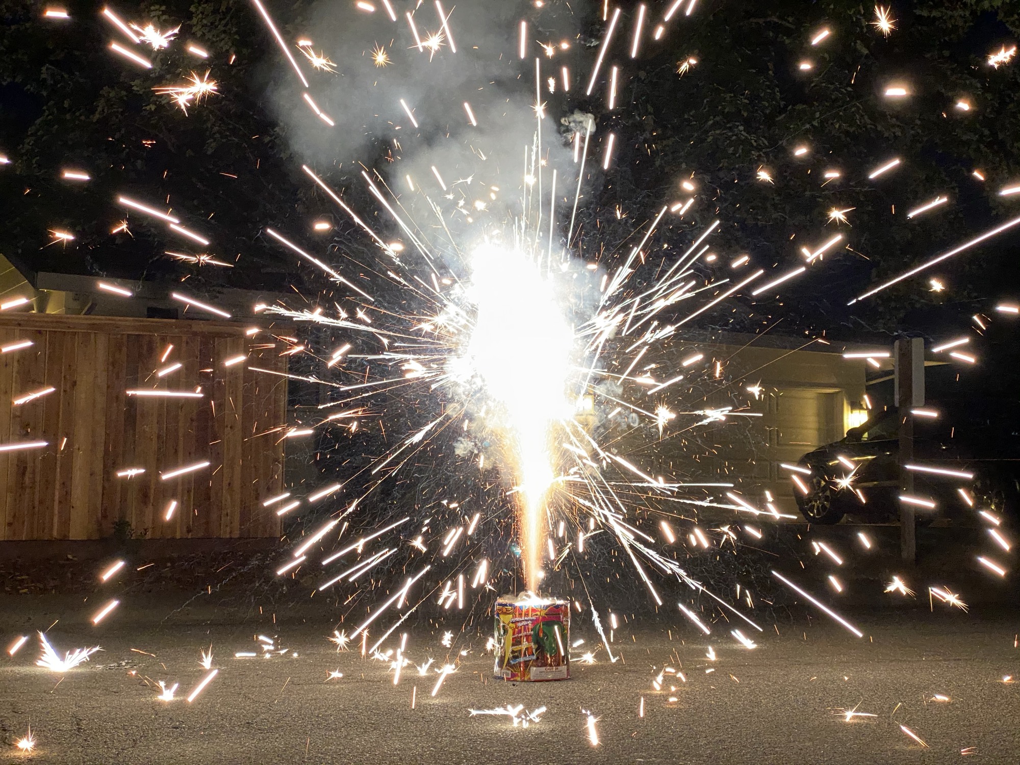 A ground firework fountain shoots bright white and orange sparks in all directions on a paved driveway at night. In the background, a wooden fence, a garage, and a parked car are visible under the cover of darkness.
