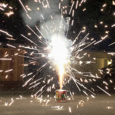 A ground firework fountain shoots bright white and orange sparks in all directions on a paved driveway at night. In the background, a wooden fence, a garage, and a parked car are visible under the cover of darkness.