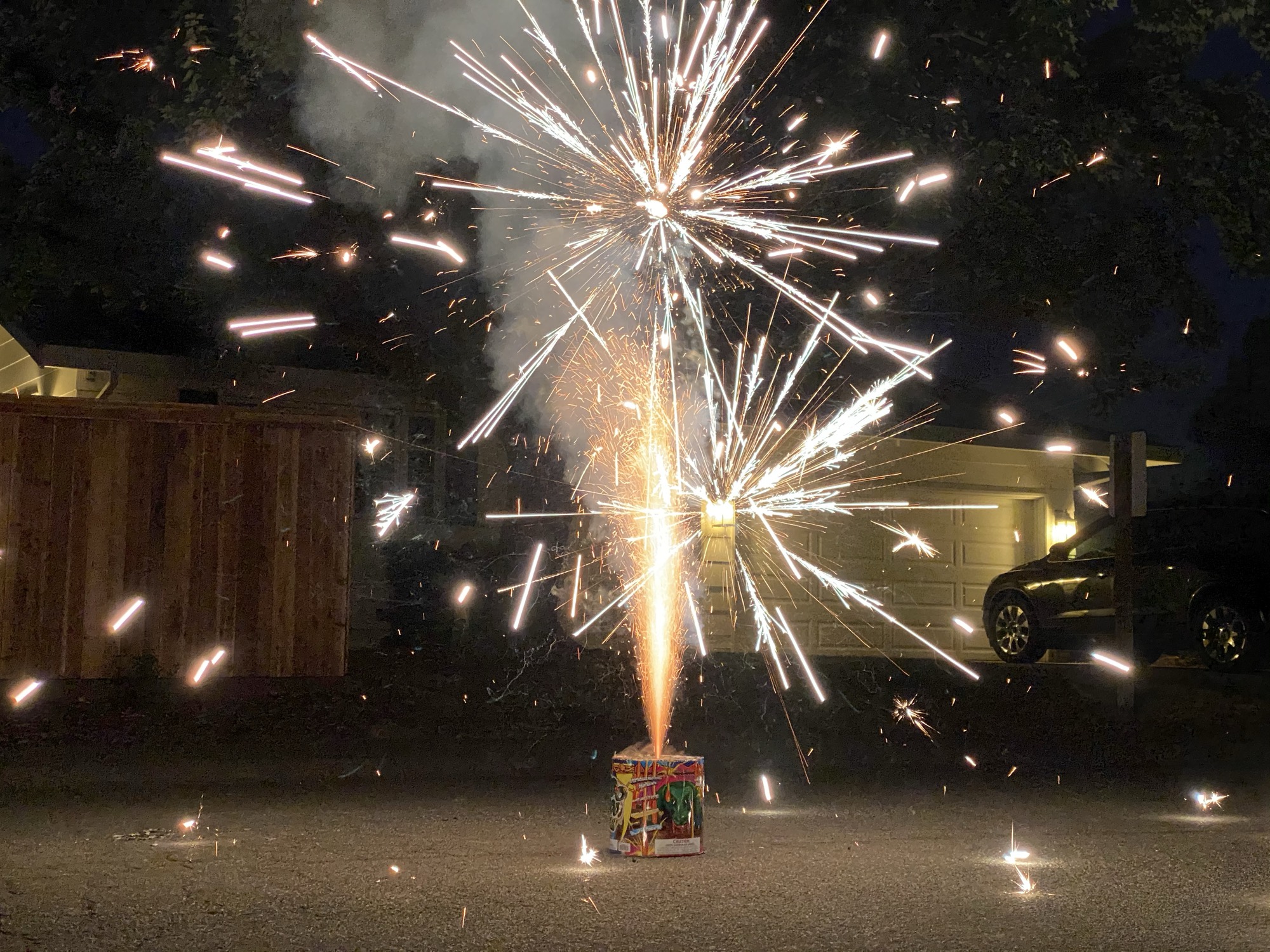 A fountain firework sits on a paved driveway at night, shooting bright white and orange sparks and smoke into the air. In the background, a residential setting is visible, featuring a wooden fence, a house with a garage door, and a parked car.