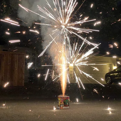 A fountain firework sits on a paved driveway at night, shooting bright white and orange sparks and smoke into the air. In the background, a residential setting is visible, featuring a wooden fence, a house with a garage door, and a parked car.