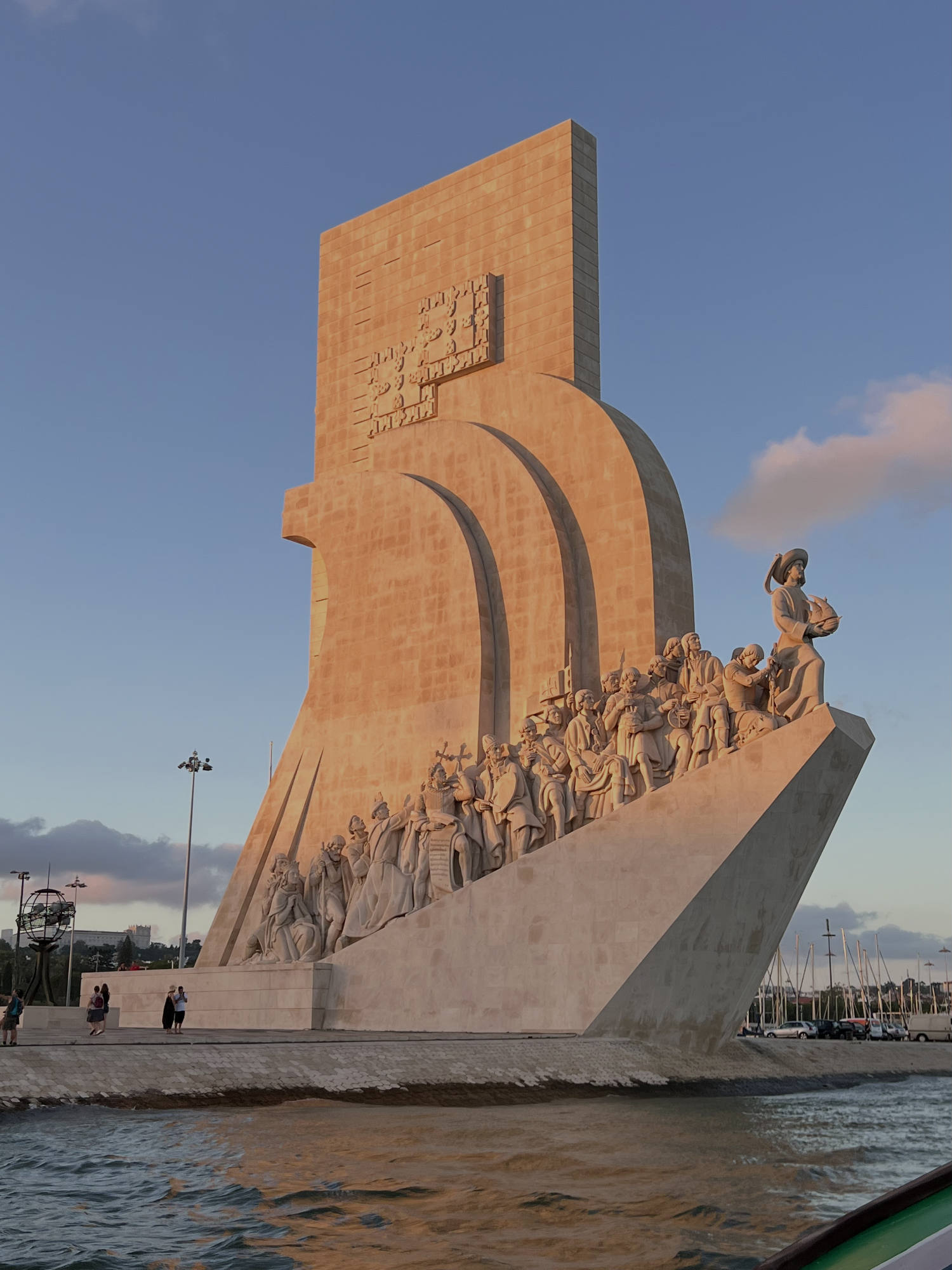 This photo captures the Monument to the Discoveries in Lisbon, Portugal, illuminated by the warm light of a setting sun. The large stone monument, shaped like a ship's prow with numerous historical figures, stands on the waterfront of the Tagus River under a blue sky.