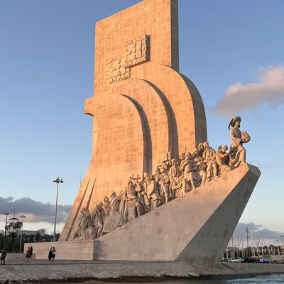 This photo captures the Monument to the Discoveries in Lisbon, Portugal, illuminated by the warm light of a setting sun. The large stone monument, shaped like a ship's prow with numerous historical figures, stands on the waterfront of the Tagus River under a blue sky.