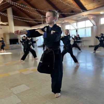 A young boy in a black martial arts uniform and brown belt practices a stance on one leg in the foreground of a spacious indoor hall with a vaulted wooden ceiling. In the background, other children in matching uniforms are also practicing martial arts poses.