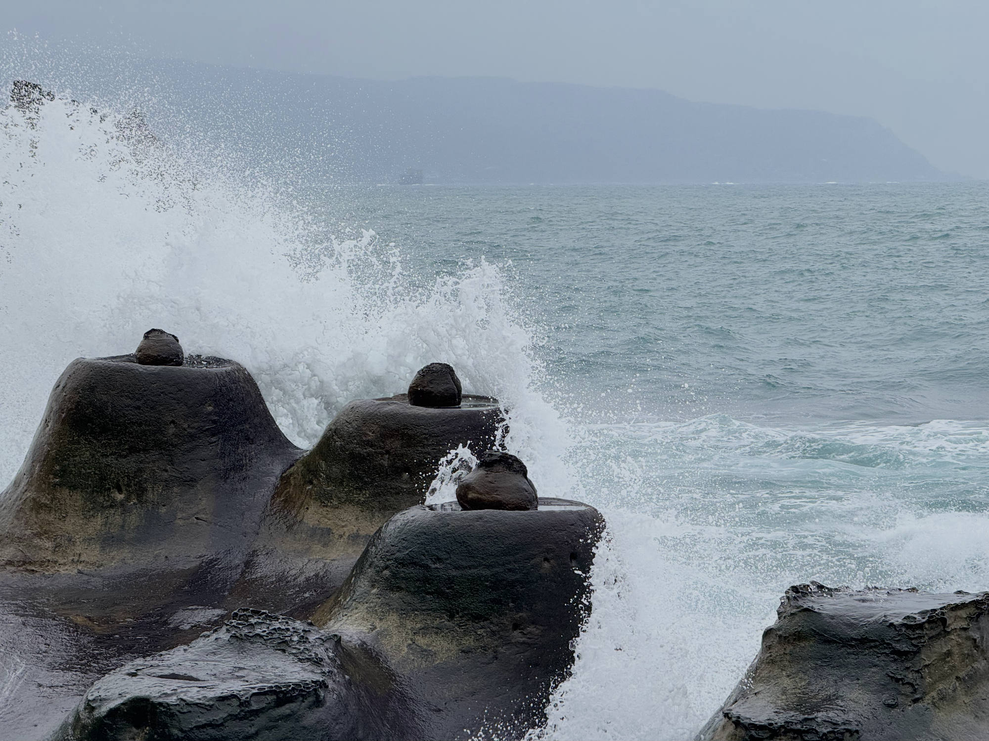 Ocean waves crash against dark, rounded rock formations along a coastline on a cloudy day. In the background, a hazy mountain range is visible across the water.