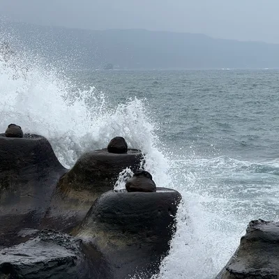 Ocean waves crash against dark, rounded rock formations along a coastline on a cloudy day. In the background, a hazy mountain range is visible across the water.