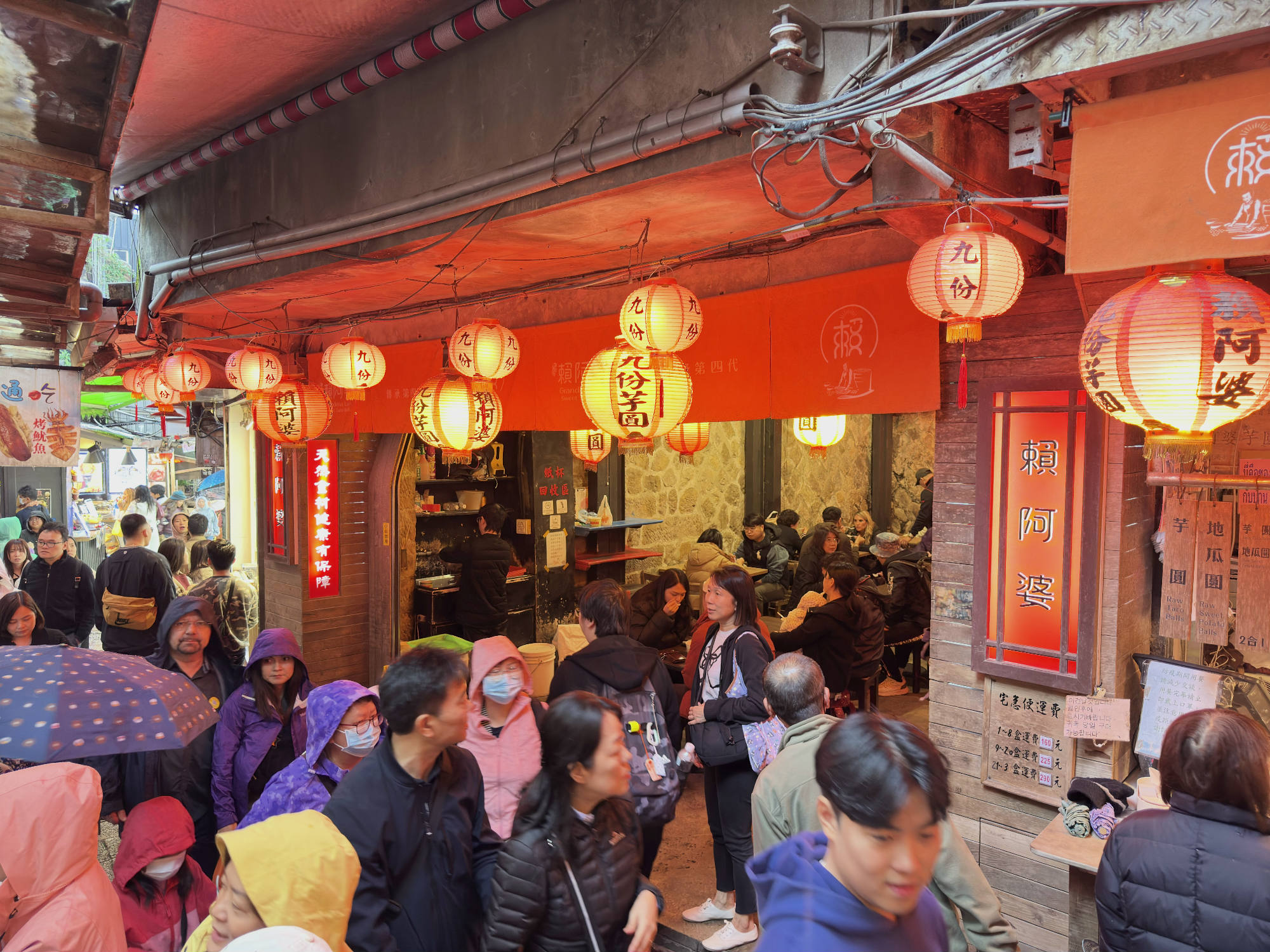 A crowd of people, some wearing rain gear and carrying umbrellas, walks along a narrow, covered street lined with shops in Jiufen, Taiwan. Red paper lanterns hang overhead, illuminating the bustling alleyway and the patrons dining inside a restaurant.