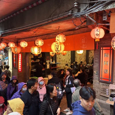 A crowd of people, some wearing rain gear and carrying umbrellas, walks along a narrow, covered street lined with shops in Jiufen, Taiwan. Red paper lanterns hang overhead, illuminating the bustling alleyway and the patrons dining inside a restaurant.
